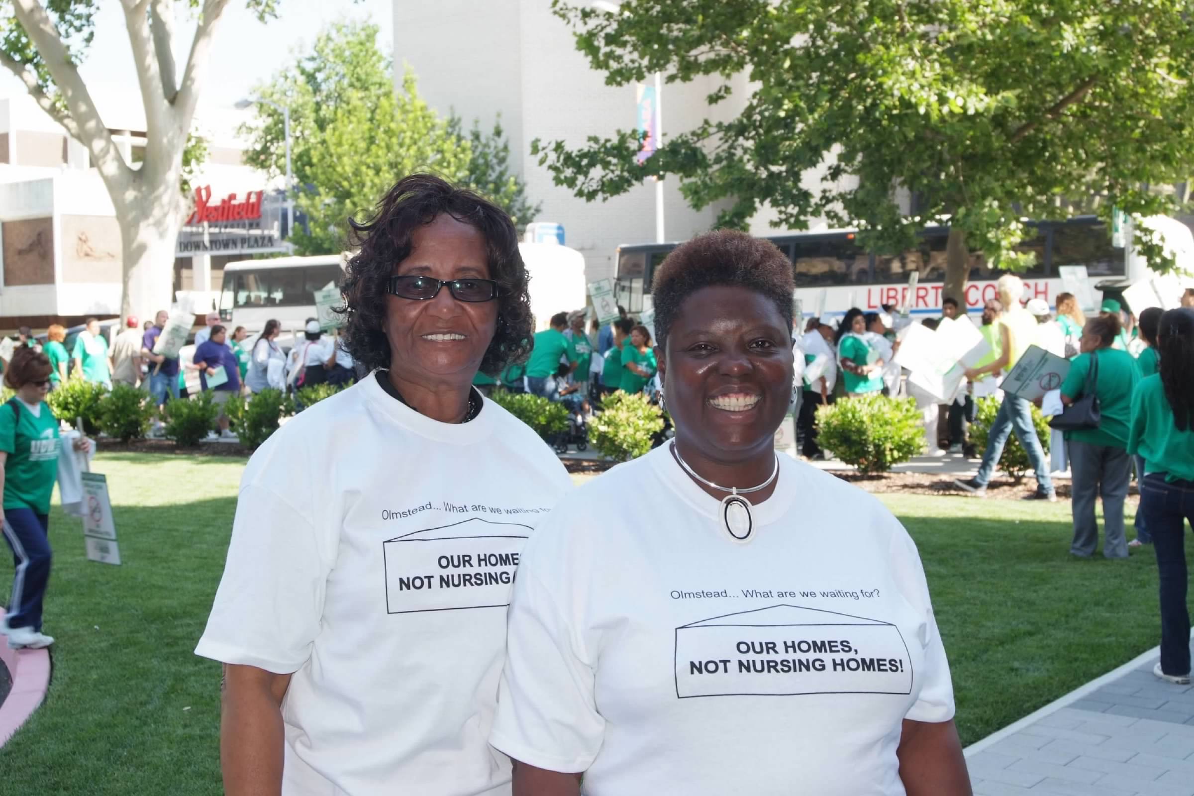 Photo of two Black women smiling for the camera in front of a grass field with people organizing. The women are wearing "our homes, not nursing homes" white t-shirts.