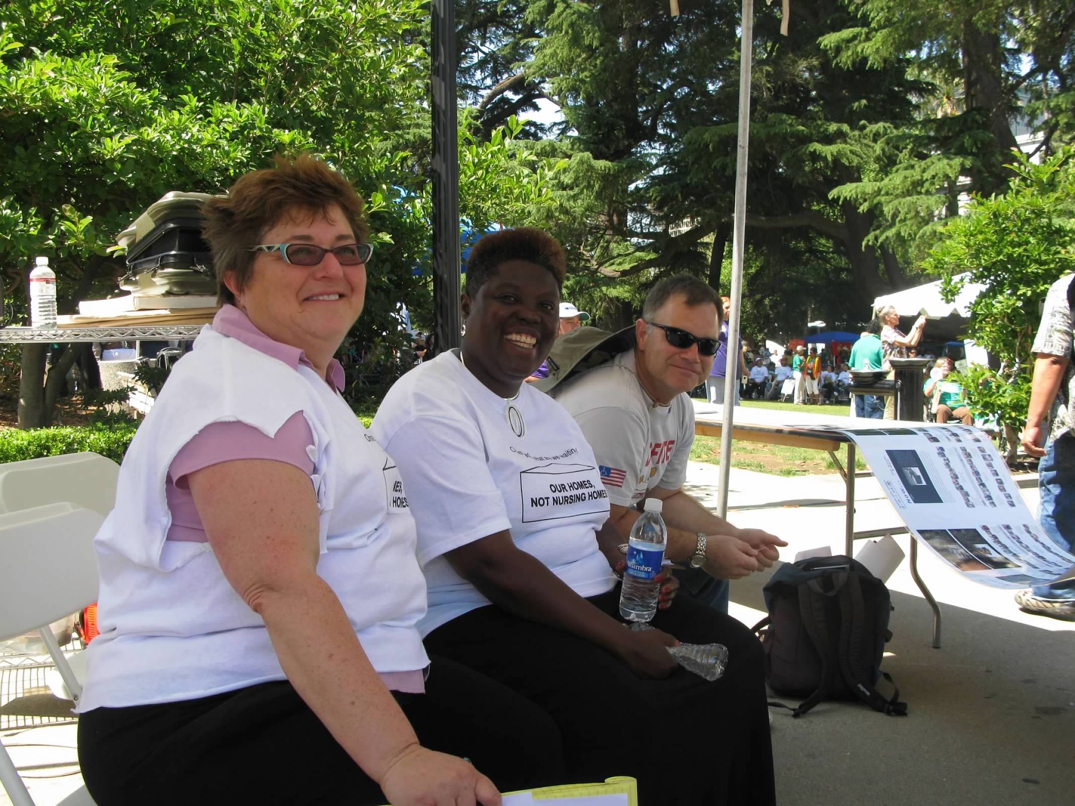 Photo of three individuals sitting together in a shaded area. From left to right: a white woman with brown hair and blue square glasses, a black woman with dangling earrings holding a water bottle, and a white man with his hands resting on his thighs and a bucket hat hanging by his neck. Lois with former DRC Lobbyist, Deborah Doctor and DAC Executive Director, Evan LeVang.
